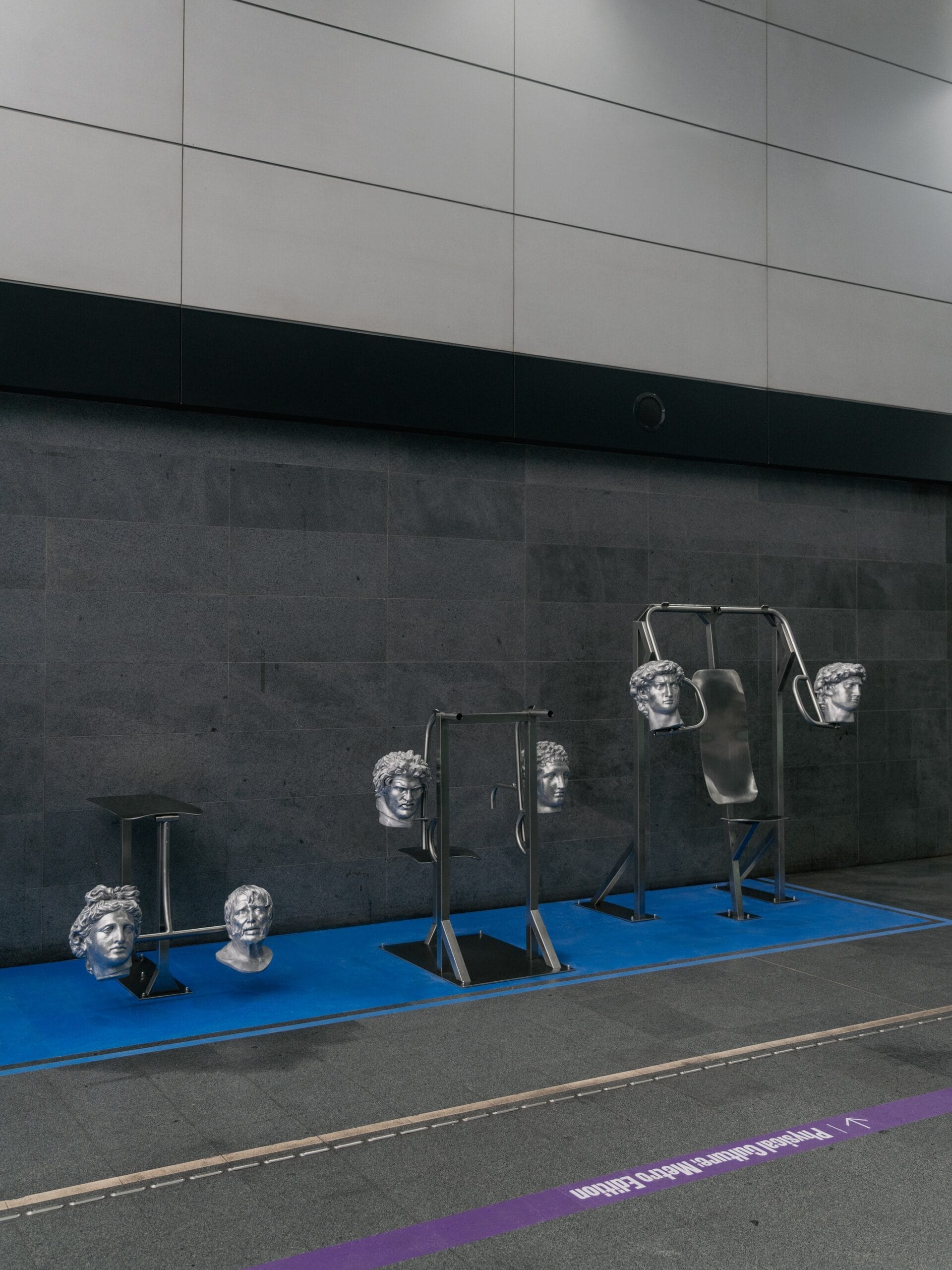 Physical Culture: Metro Edition: attrezzi da palestra in acciaio con teste classiche argentate su una pedana blu, lungo la parete scura della stazione